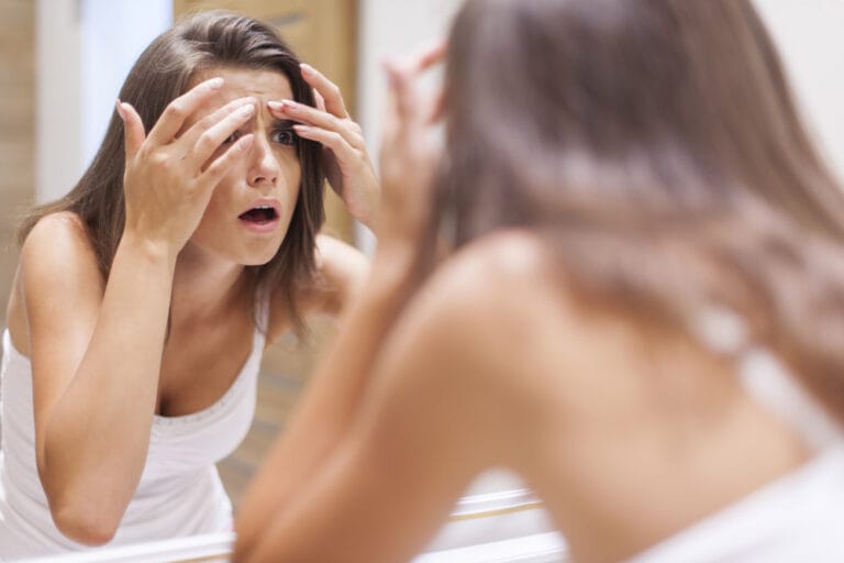 Junge Frau schaut schockiert in den Spiegel auf ihre Zornesfalte