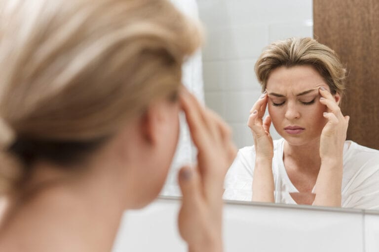 Frau steht vor dem Spiegel und hält sich die Schläfen wegen Migräne