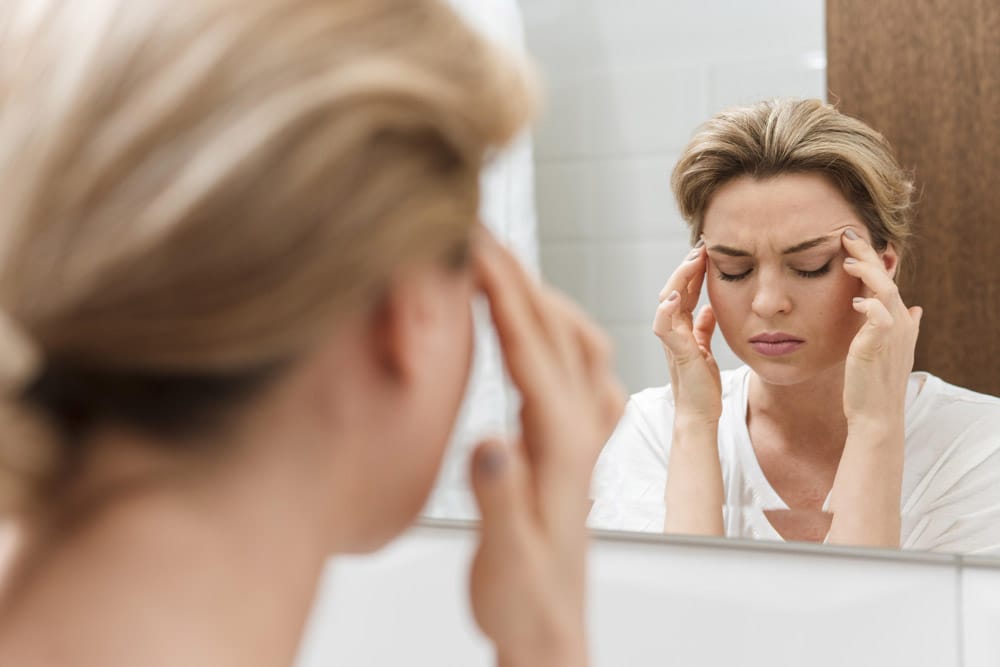 Frau steht vor dem Spiegel und hält sich die Schläfen wegen Migräne